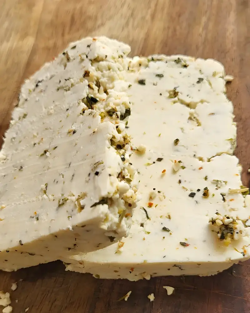 A cut block of PANEVO Oregano paneer on a wooden board, flecks of green oregano through the cut face